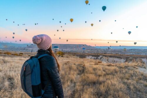 Woman watches hot air balloons rise over a dry, scenic valley at sunrise.